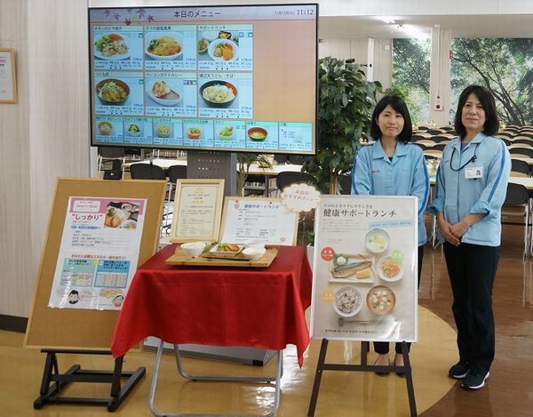 食堂のメニュー掲示と、ヤマハ発動機ビズパートナー 名倉麻美さん(左)・前田直美さん(右)